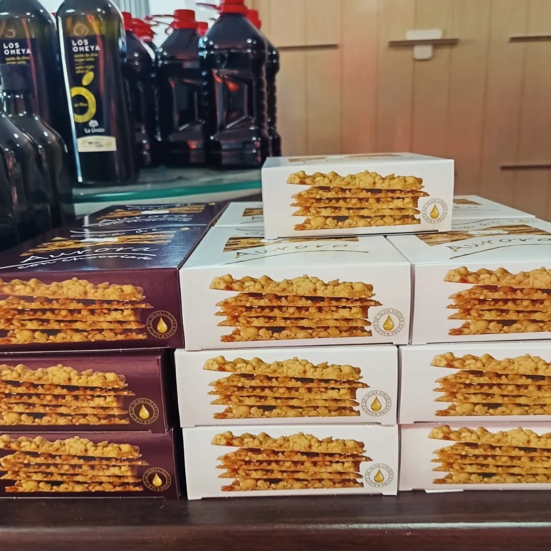 coca-de-aurora-cuadrada-1-min Primer plano de una pila ordenada de cajas rectangulares de un dulce llamado “Coca de Aurora”. Las cajas son principalmente blancas y burdeos, y muestran en la parte frontal la fotografía de una coca cuadrada formada por varias capas finas de masa crujiente con migas doradas por encima. A la izquierda y al fondo se ven varias botellas altas y oscuras con tapones rojos, colocadas en una estantería o mostrador de tienda. El ambiente es el de una tienda de alimentación o producto gourmet.