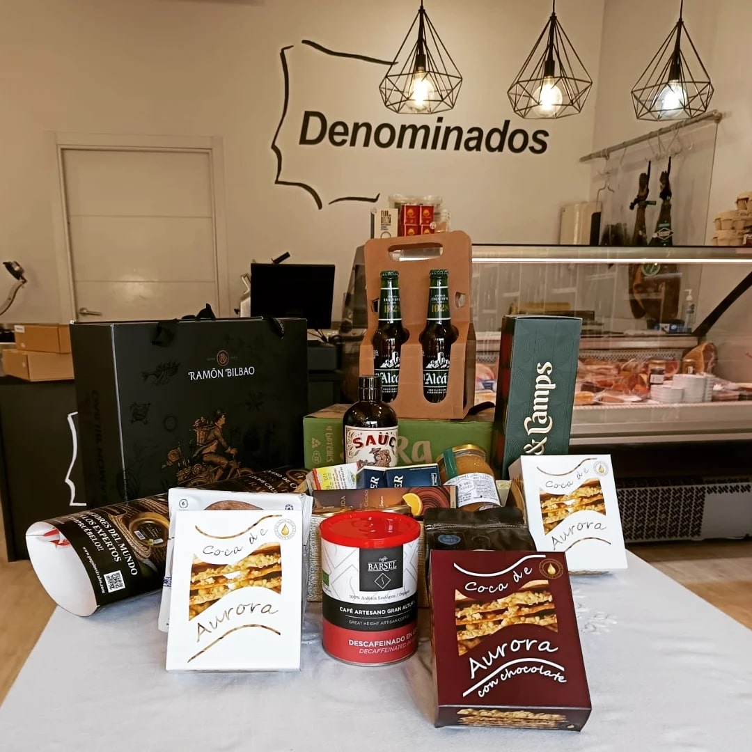 coca-de-aurora-cuadrada-mostrador1-min En el centro de la imagen hay una mesa cubierta con un mantel claro sobre la que se ha dispuesto un lote de productos gastronómicos: varias cajas de Coca de Aurora (blancas y burdeos), una lata cilíndrica de café con tapa roja, botellas de cerveza en un soporte de cartón, una botella de salsa, paquetes de café y otros artículos. Al fondo se ve el interior de una tienda delicatessen: un mostrador refrigerado con embutidos o productos cárnicos, una bolsa de regalo negra con el logo “Ramón Bilbao” a la izquierda y, en la pared del fondo, el rótulo grande que dice “Denominados” con una silueta de mapa alrededor. La iluminación es cálida y resalta el carácter de tienda gourmet.