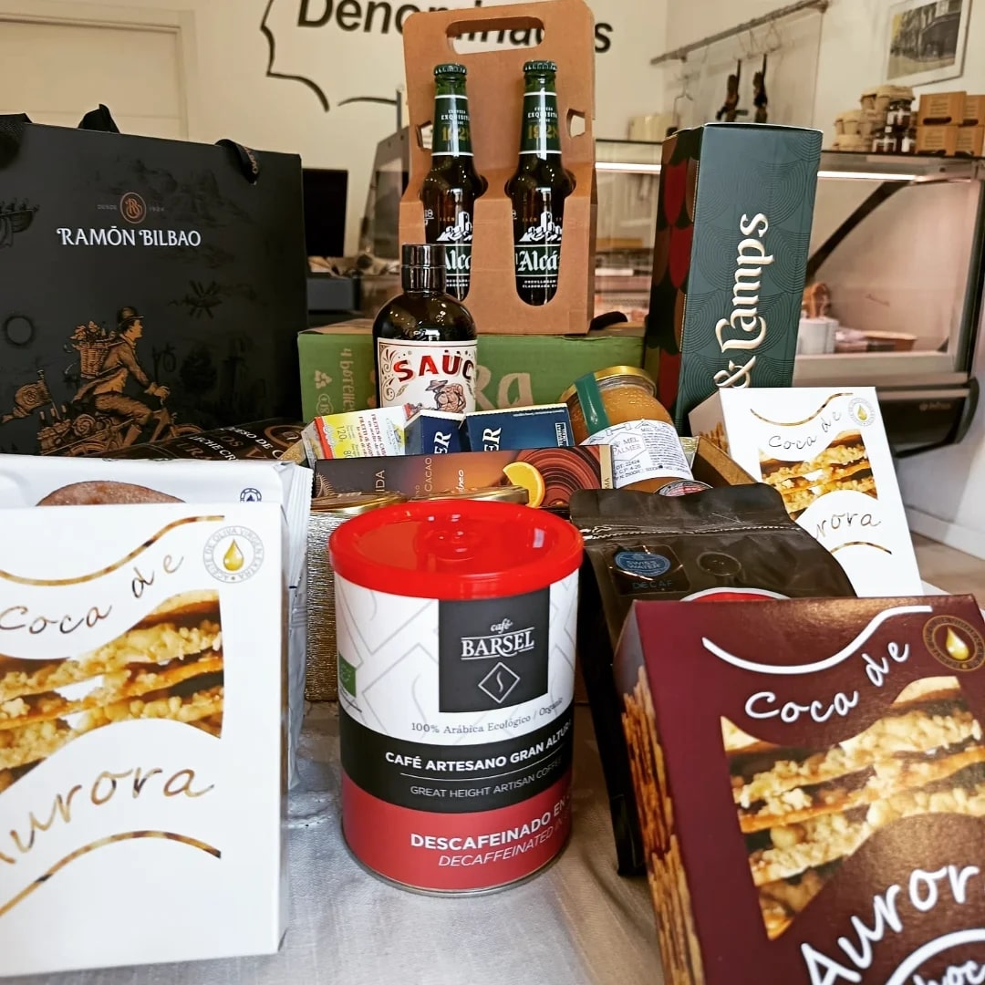 coca-de-aurora-cuadrada-mostrador2-min La imagen se centra en la parte frontal del lote de productos. En primer plano se ven varias cajas de Coca de Aurora (una blanca y otra burdeos) y, en el centro, una lata grande de café artesanal Barcelo con tapa roja. A su alrededor hay otros productos: paquetes de café, botes, una botella de salsa, un estuche verde de vino o licor y un pack de dos cervezas en un soporte de cartón. Al fondo, ligeramente desenfocado, se distingue el mostrador de la tienda y parte del rótulo “Denominados” en la pared. La composición destaca la Coca de Aurora como parte de un conjunto de regalos o productos gourmet.